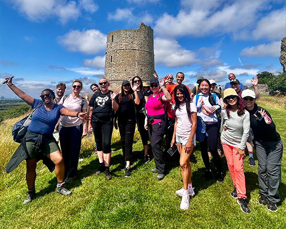 A diverse group of people posing happily outdoors near a historical tower on a sunny day showcasing teamwork and camaraderie in their 5 hiking adventure.